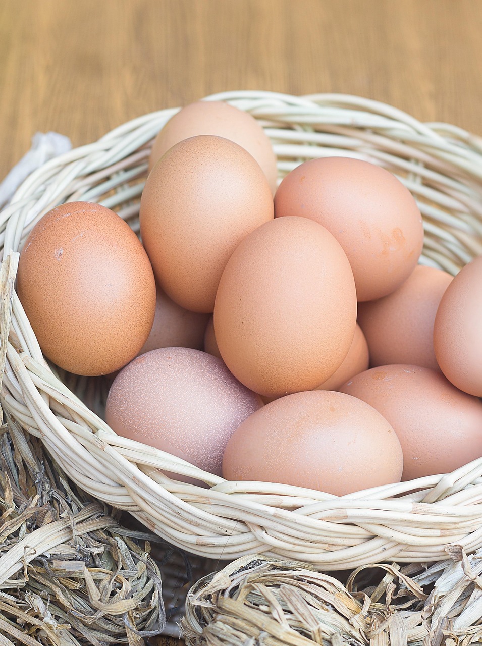 A wicker basket filled with fresh brown eggs sitting on a rustic wooden surface, surrounded by dried corn husks.