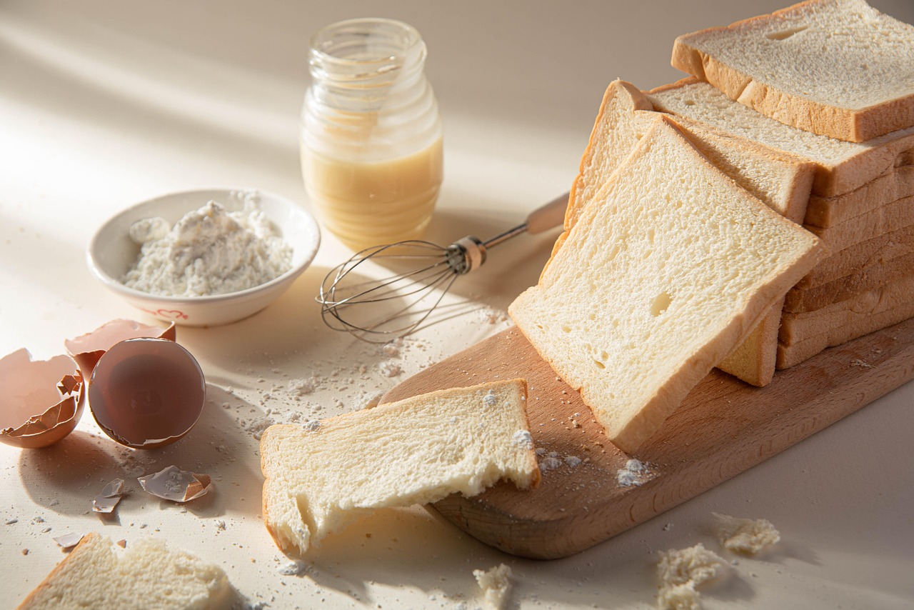 Slices of soft, homemade sandwich bread on a wooden cutting board surrounded by baking ingredients like flour, eggshells, and a whisk.