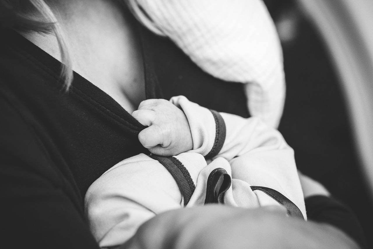 Black and white close-up of a baby nursing, gently grasping their mother’s shirt during a breastfeeding session.