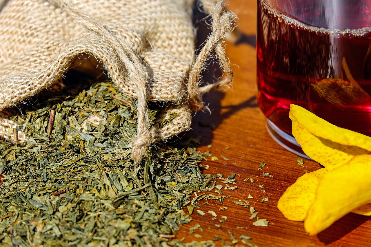 Dried herbal leaves spilling from a burlap sack next to a glass of herbal tea and yellow flower petals on a wooden surface.