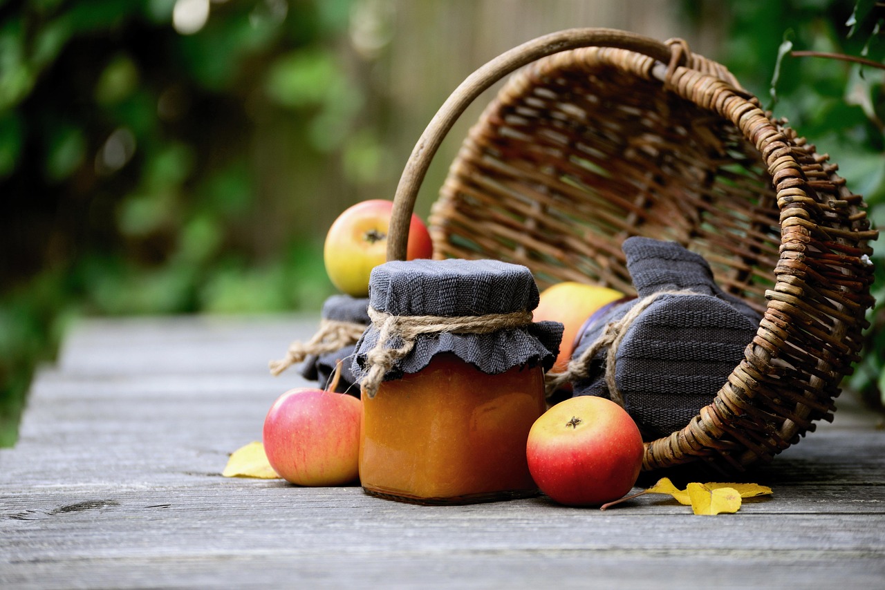 Homemade preserves in jars wrapped with cloth and twine, surrounded by apples and a tipped-over wicker basket on a rustic wooden table.