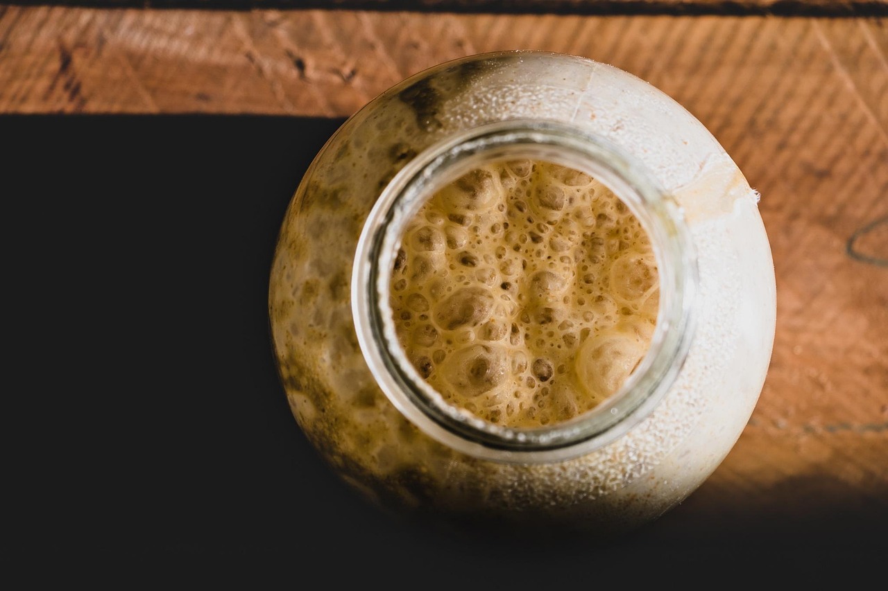 Overhead view of a bubbly, active sourdough starter in a glass jar on a wooden surface, showing natural fermentation in progress.