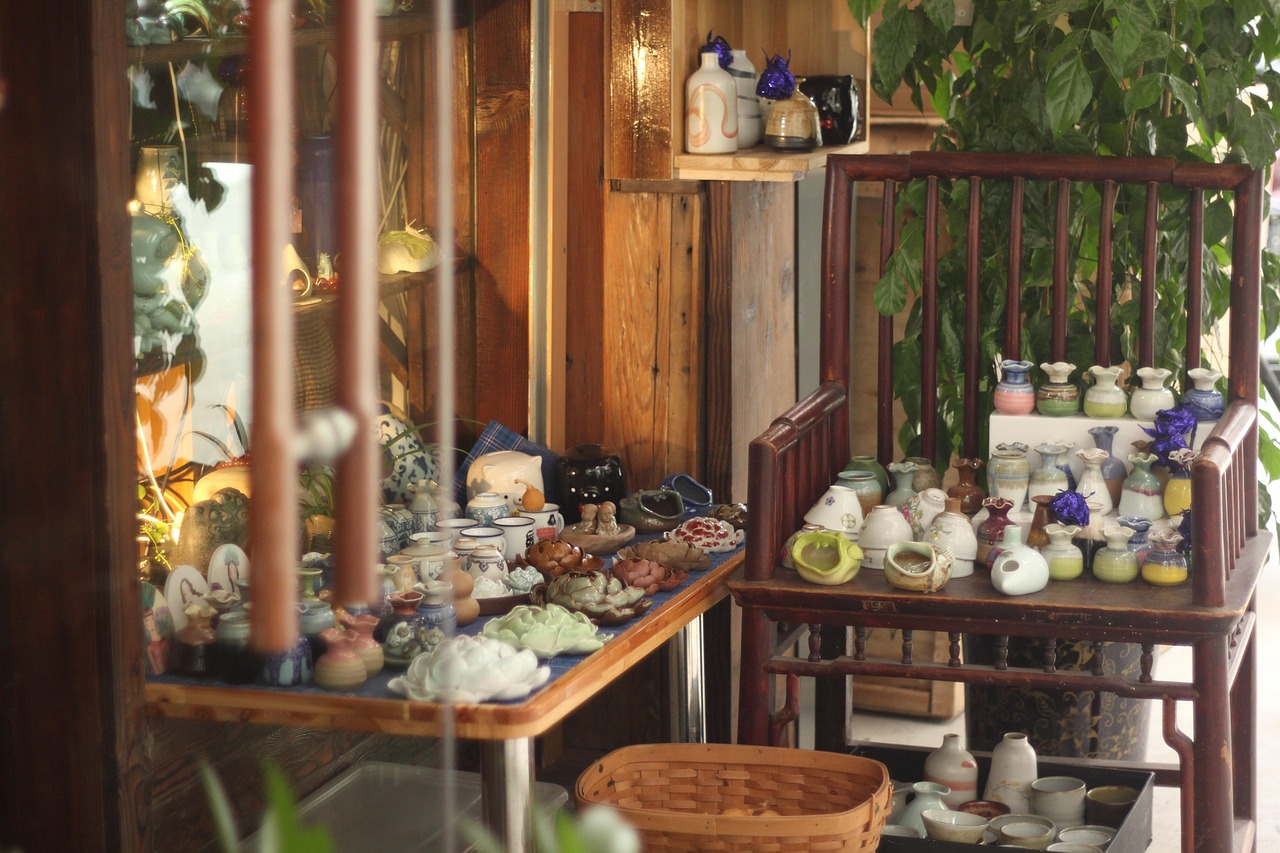 Display of assorted vintage pottery, ceramic vases, and unique dishware arranged on tables and a wooden chair in a cozy thrift store corner.